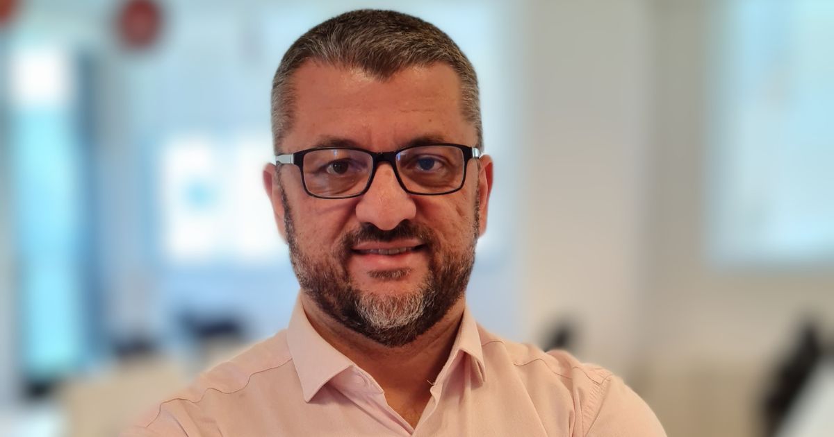 Homem de meia-idade com barba e cabelo curto, ambos grisalhos, usando óculos de armação preta e camisa social rosa clara. Ele está sorrindo levemente e olhando diretamente para a câmera. O fundo está desfocado, sugerindo um ambiente de escritório moderno e bem iluminado, com móveis e janelas visíveis de forma indistinta.