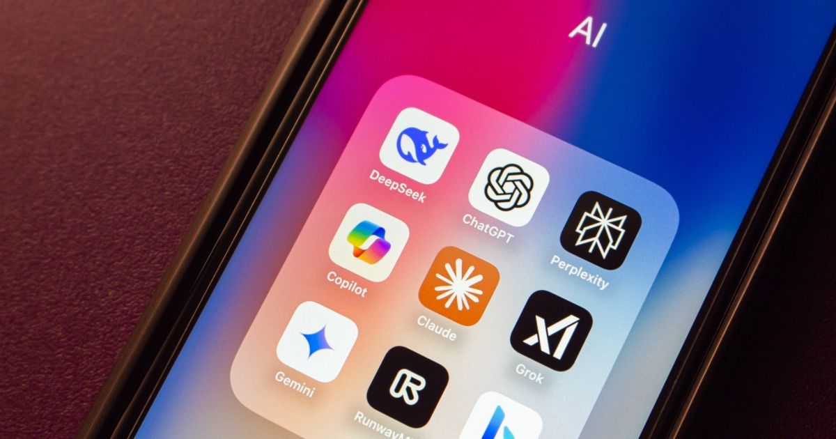 Ecrã de um smartphone com uma pasta de aplicações intitulada 'AI', contendo ícones de várias plataformas de inteligência artificial. São visíveis nove aplicações: DeepSeek (ícone de baleia azul), ChatGPT (ícone com um nó hexagonal entrelaçado em preto e branco), Perplexity (estrela geométrica branca sobre fundo preto), Copilot (ícone multicolorido em forma de cruz), Claude (ícone laranja com traços brancos), Grok (ícone preto com um 'X' branco), Gemini (estrela azul sobre fundo branco), RunwayML (ícone preto com letra 'R' estilizada, produtos digitais) e outra app com ícone azul-claro no canto inferior direito. O fundo do ecrã é um gradiente rosa, roxo e azul.