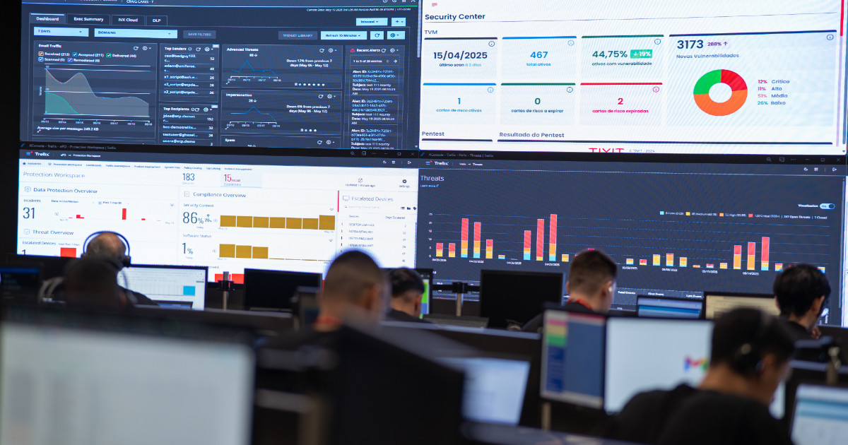 Imagem de um Centro de Operações de Segurança (SOC) da empresa Tivit, com vários profissionais concentrados em suas estações de trabalho, todos voltados para grandes painéis de monitoramento na parede. Os painéis exibem dashboards com gráficos, indicadores de segurança cibernética, tráfego de e-mails, vulnerabilidades detectadas e resultados de compliance. A imagem transmite um ambiente de vigilância tecnológica intensa, voltado à proteção de dados e gerenciamento de ameaças digitais. (Cibersegurança)