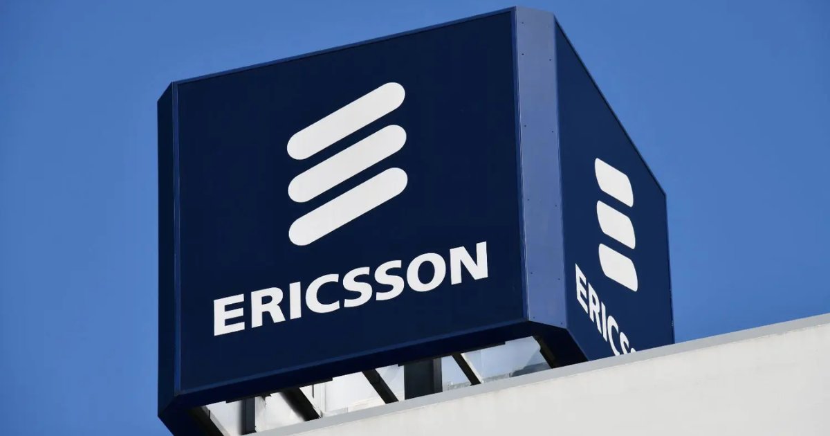 Placa com o logotipo da empresa Ericsson em destaque. O letreiro é azul-escuro com o nome "ERICSSON" em letras brancas, acompanhado do símbolo icônico da marca — três linhas brancas inclinadas paralelas. A placa está posicionada no topo de um edifício, com céu azul ao fundo (ações)