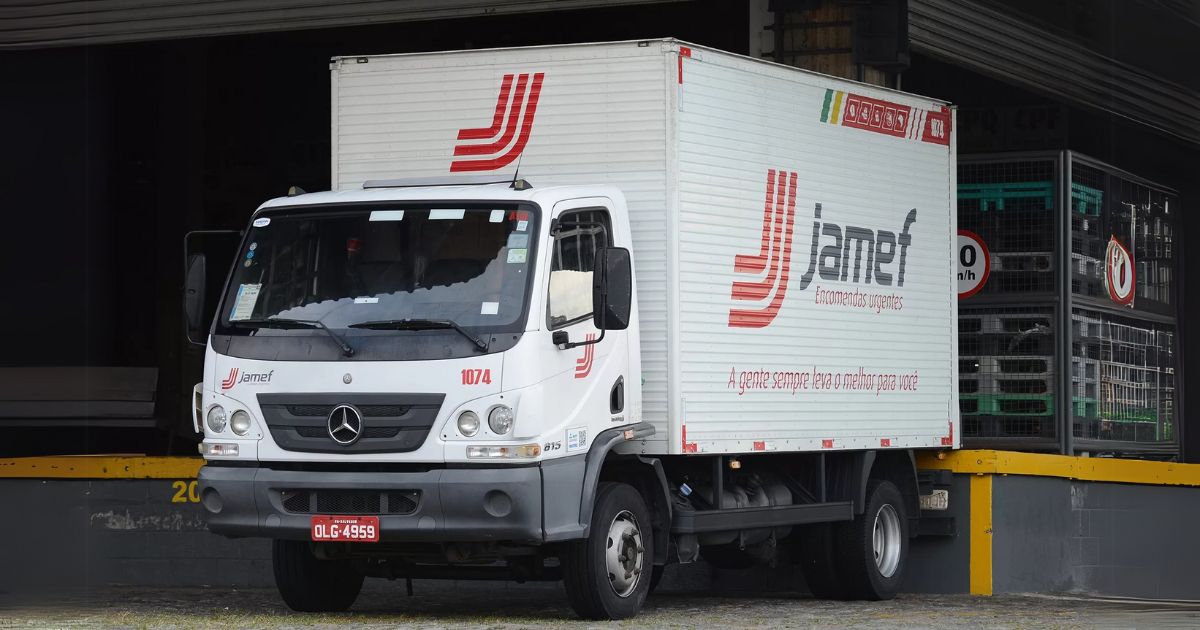 Caminhão branco da empresa Jamef, especializada em encomendas urgentes, estacionado em uma doca de carregamento. O veículo é da marca Mercedes-Benz e possui o número de identificação 1074 na lateral. A carroceria exibe o logotipo da empresa e o slogan "A gente sempre leva o melhor para você". Ao fundo, há sinalizações e estruturas metálicas com caixas organizadoras (acidentes)
