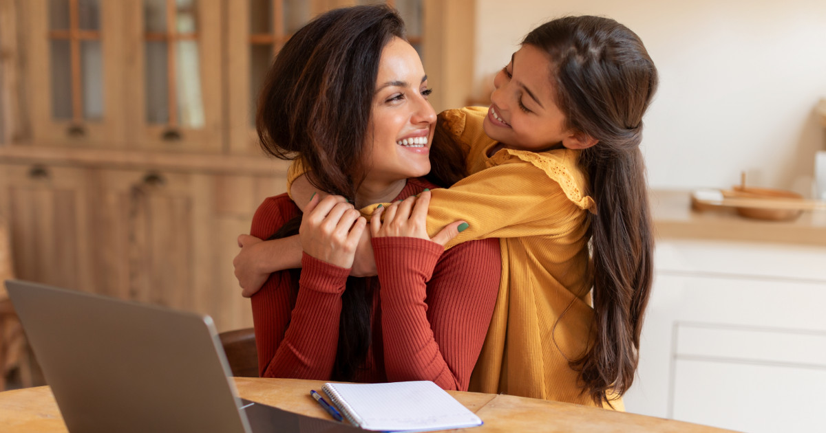 Mulher sorridente sentada à mesa de madeira com um notebook e um caderno à sua frente, sendo abraçada por uma menina que está em pé atrás dela. As duas estão trocando olhares afetuosos em um ambiente doméstico aconchegante, sugerindo uma cena de carinho entre mãe e filha durante o trabalho em casa (carreira)