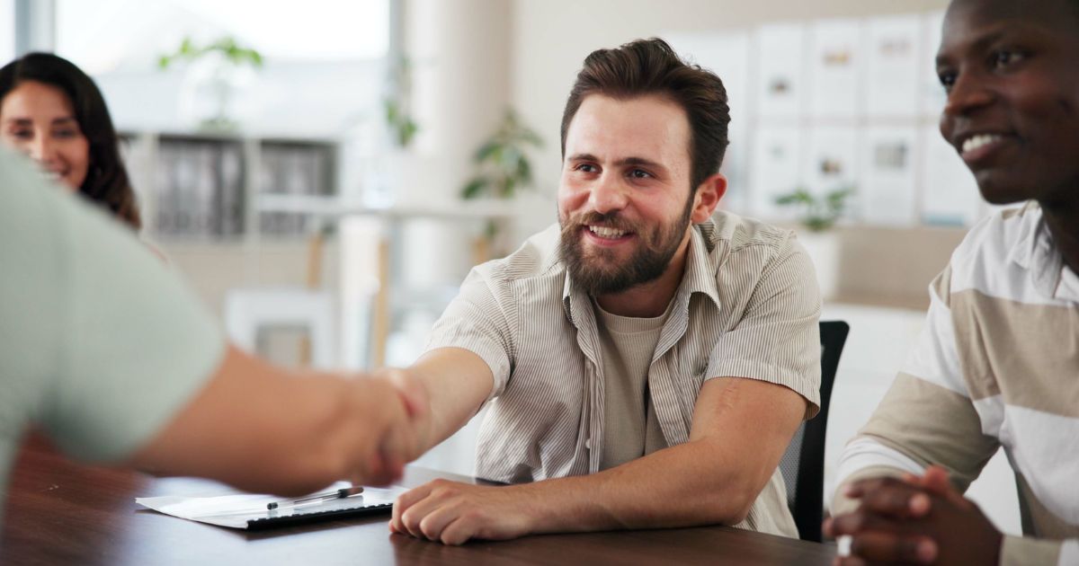 Um grupo de pessoas está sentado ao redor de uma mesa, em um ambiente que parece ser um escritório ou sala de reuniões. Uma pessoa estende a mão para cumprimentar outra, cuja mão aparece em primeiro plano. Ao fundo, há prateleiras e algumas plantas, compondo um cenário corporativo ou profissional. (Simetrik)
