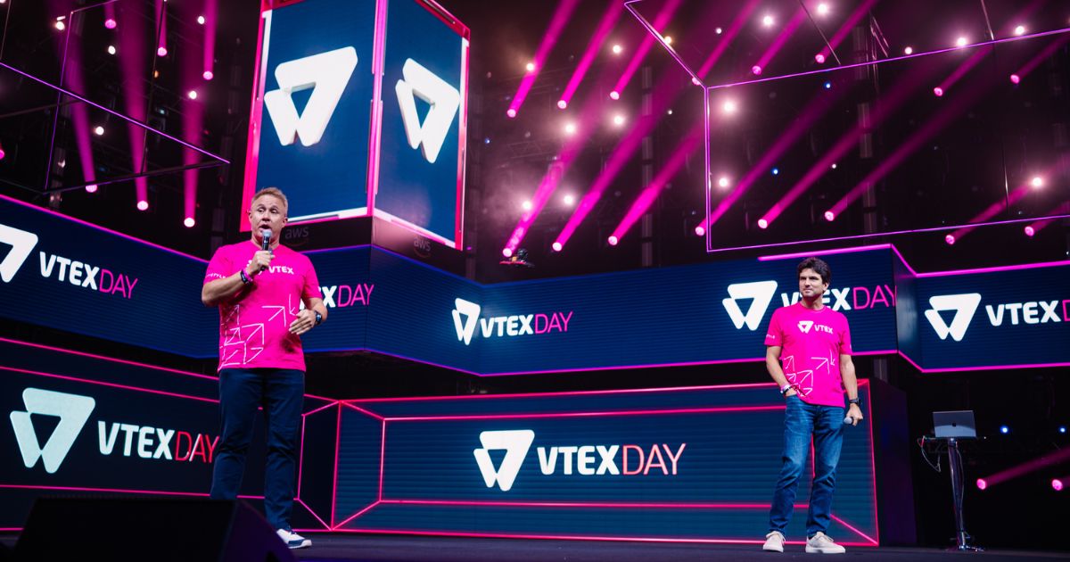 Duas pessoas estão em um palco iluminado por luzes rosa e roxas, com várias telas ao fundo exibindo o texto "VTEX DAY" e o logotipo da VTEX. Ambas vestem camisetas rosa com o logotipo da VTEX e padrões geométricos. O ambiente transmite uma atmosfera vibrante e tecnológica, típica de eventos corporativos ou conferências de inovação.