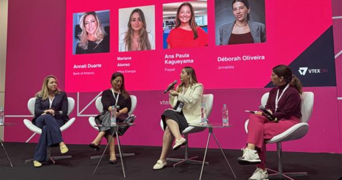 Quatro mulheres participam de um painel de discussão em um palco. Cada uma está sentada com um microfone em mãos, envolvidas em uma conversa sobre transformação digital. Ao fundo, uma grande tela exibe seus nomes e respectivas organizações: Annali Duarte (Bank of America), Mariana Alonso (Votorantim Energia), Ana Paula Kagueyama (Paypal) e Déborah Oliveira (Jornalista). O logotipo da VTEX na tela indica que o evento é organizado por essa empresa.