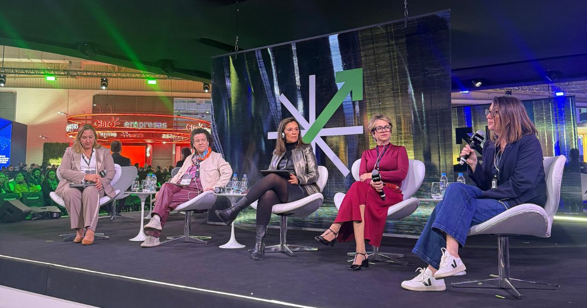 Cinco mulheres sentadas em cadeiras brancas no palco de um evento de tecnologia. Elas participam de um painel sobre diversidade e inteligência artificial, com um telão ao fundo exibindo a imagem ampliada da moderadora falando ao microfone. O ambiente é moderno, com iluminação cênica e público ao fundo. Foto: Déborah Oliveira