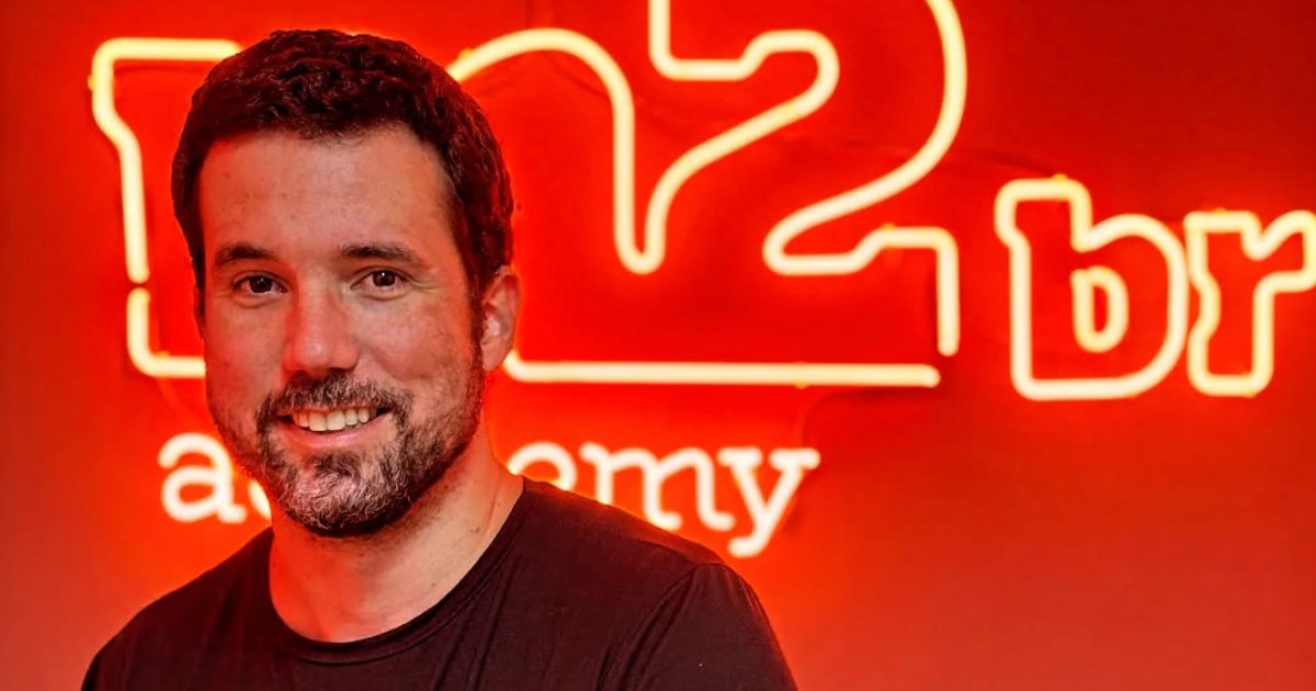 Imagem de Ricardo Marsili, especialista em inteligência artificial, sorrindo em frente a uma parede vermelha iluminada com letreiro em neon da M2BR Academy. Ele veste uma camiseta preta e está em um ambiente com iluminação quente, destacando o logotipo ao fundo. (Ricardo Marsili, M2BR, Brasil)