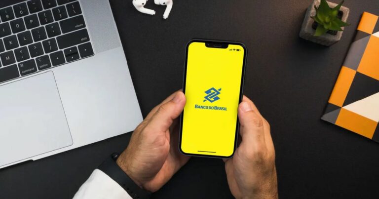Imagem de uma pessoa segurando um smartphone com a tela exibindo o logotipo do Banco do Brasil sobre um fundo amarelo. O ambiente inclui um notebook prateado no canto superior esquerdo, um par de fones de ouvido sem fio brancos acima do smartphone, um pequeno vaso com uma planta no canto superior direito e um caderno com capa geométrica nas cores laranja, preto, branco e cinza à direita. A superfície da mesa é preta.