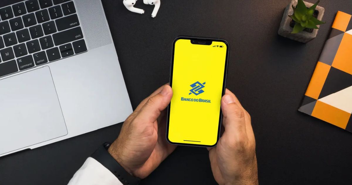 Imagem de uma pessoa segurando um smartphone com a tela exibindo o logotipo do Banco do Brasil sobre um fundo amarelo. O ambiente inclui um notebook prateado no canto superior esquerdo, um par de fones de ouvido sem fio brancos acima do smartphone, um pequeno vaso com uma planta no canto superior direito e um caderno com capa geométrica nas cores laranja, preto, branco e cinza à direita. A superfície da mesa é preta.